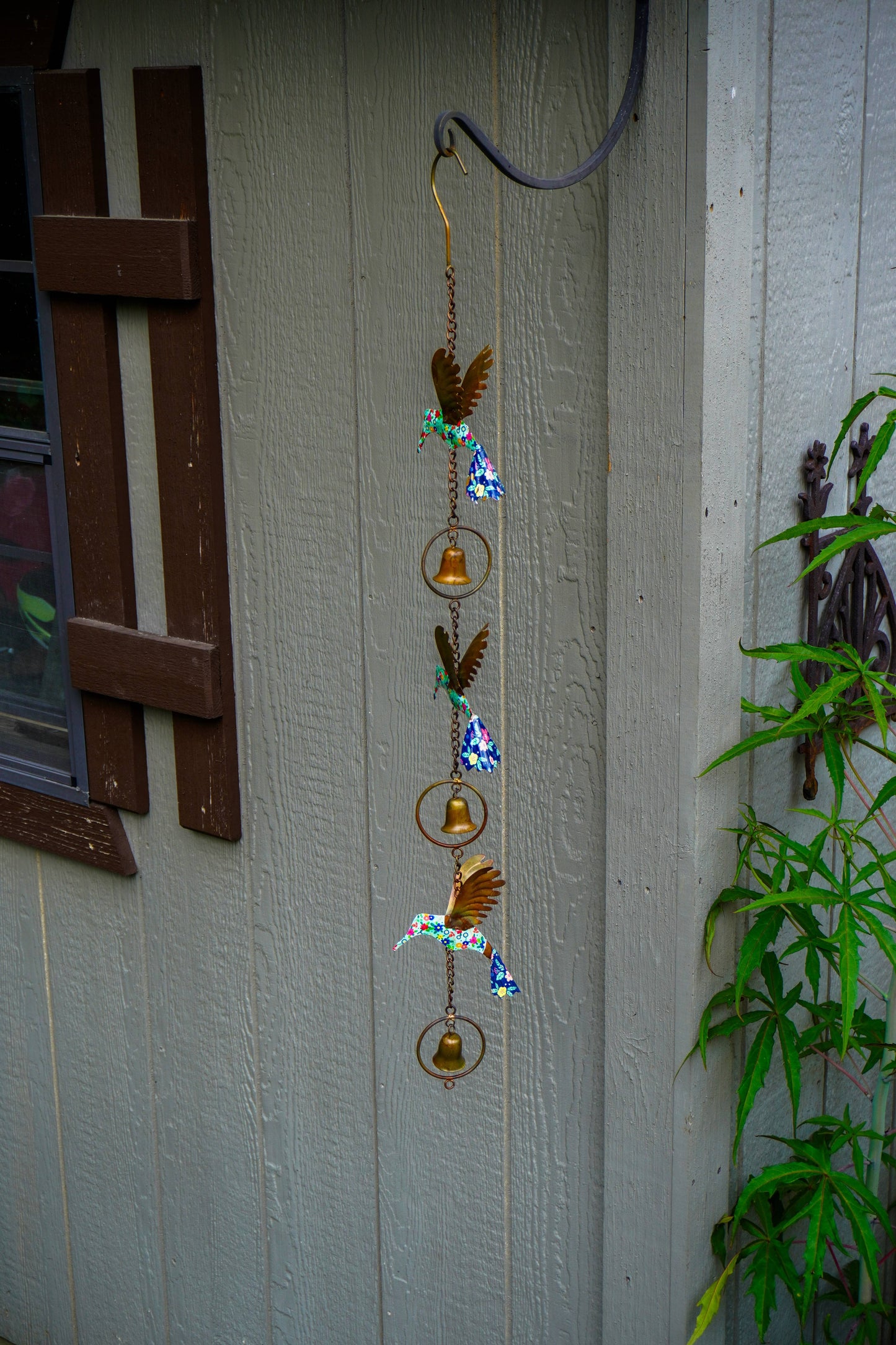 A hanging metal wind chime with bird and bell motifs, displayed outdoors against a gray wooden wall next to green plants.