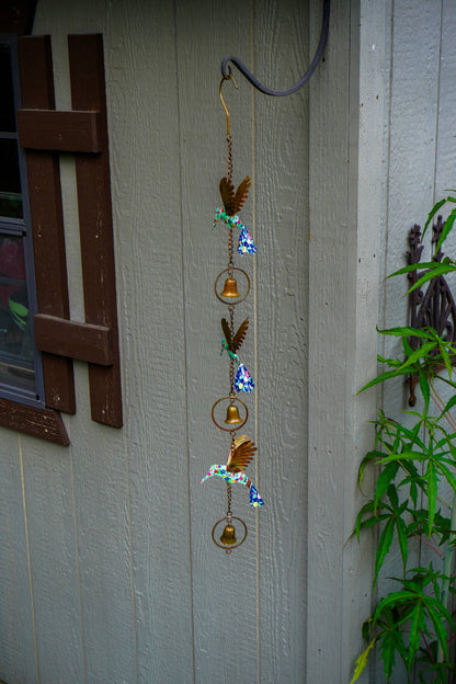 A hanging metal wind chime with bird and bell motifs, displayed outdoors against a gray wooden wall next to green plants.
