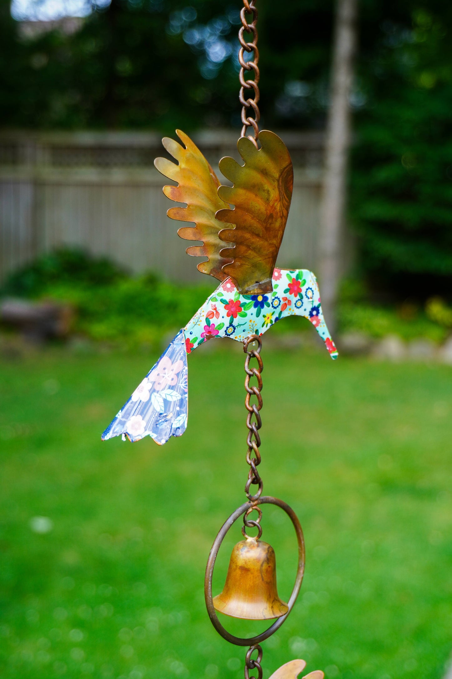 A handmade metal wind spinner shaped like a hummingbird, featuring a colorful floral-patterned body, metallic gold wings, and a small gold bell hanging below, set against a blurred green backyard background.