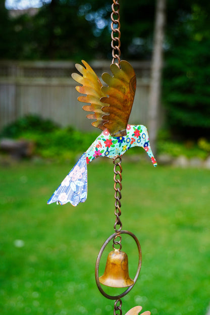 A handmade metal wind spinner shaped like a hummingbird, featuring a colorful floral-patterned body, metallic gold wings, and a small gold bell hanging below, set against a blurred green backyard background.