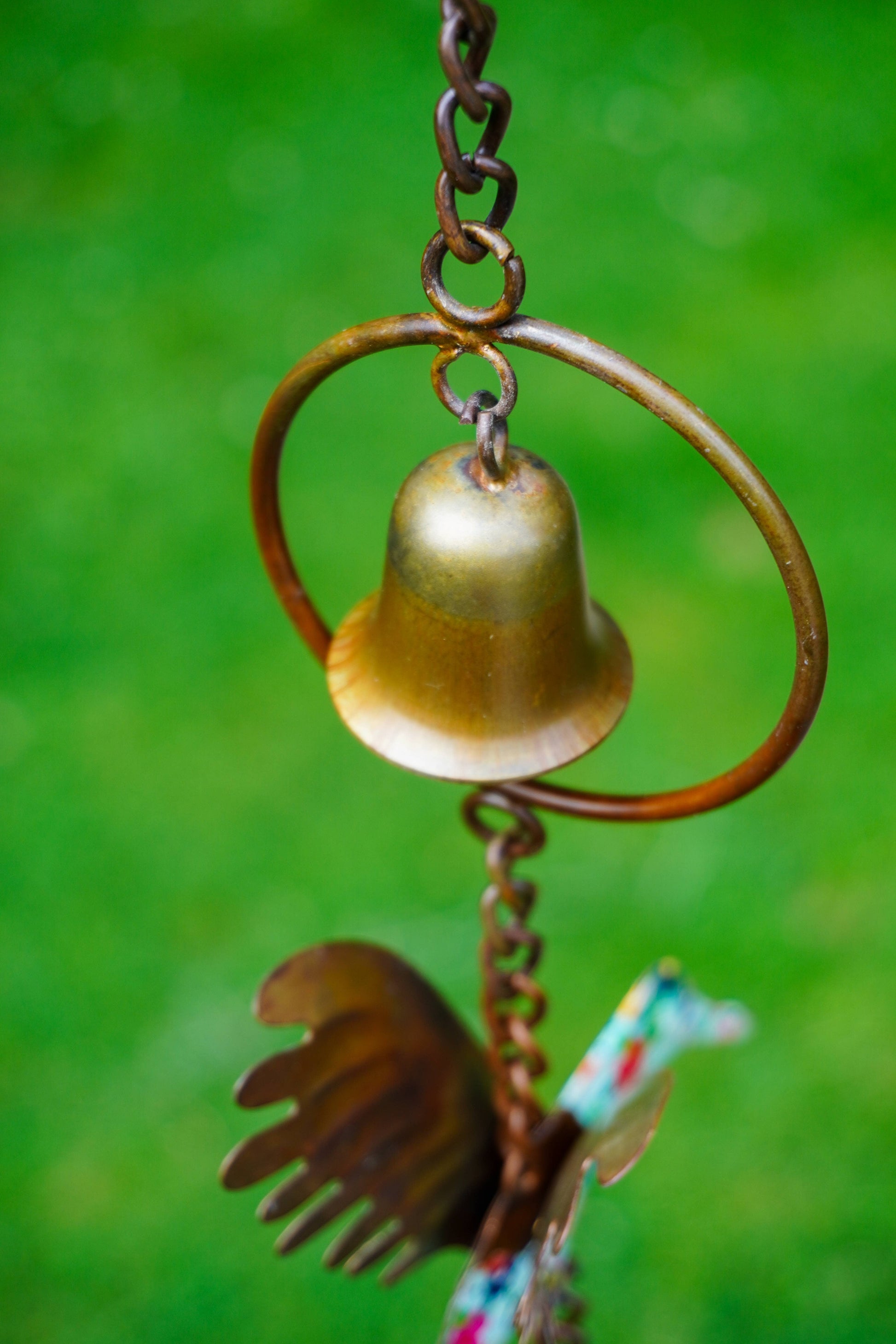 A rustic metal garden ornament hanging outdoors against a blurred green background. The ornament features a small gold-toned bell inside a circular ring, with a weathered brown bird and leaf details below.