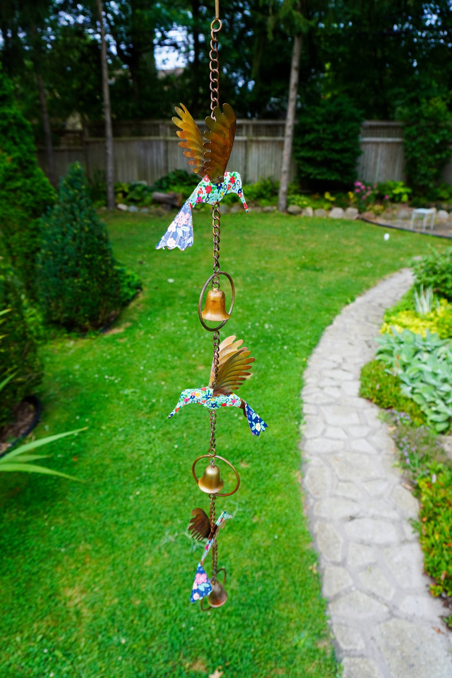 A hanging metal garden ornament with bird and bell motifs, photographed outdoors over a green lawn with a stone path and trees in the background.
