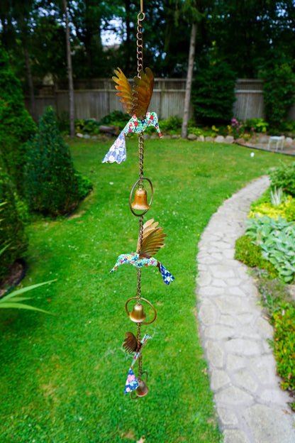 A hanging metal garden ornament with bird and bell motifs, photographed outdoors over a green lawn with a stone path and trees in the background.