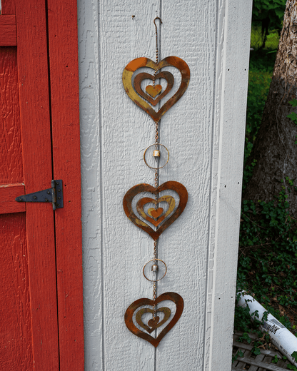 Three rustic metal heart wind chimes with concentric cutout hearts hang vertically on a white exterior wall next to a red door, with outdoor greenery and a tree trunk visible on the right.