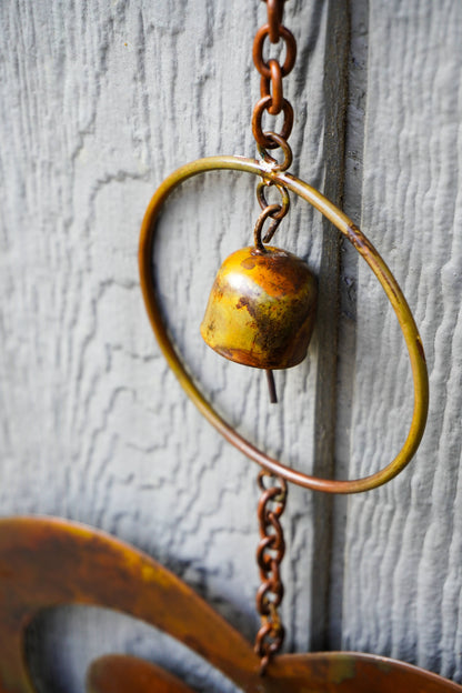 Rustic metal wind chime hanging against a weathered gray wood door, featuring a small bell suspended inside a large circular hoop with a chain.
