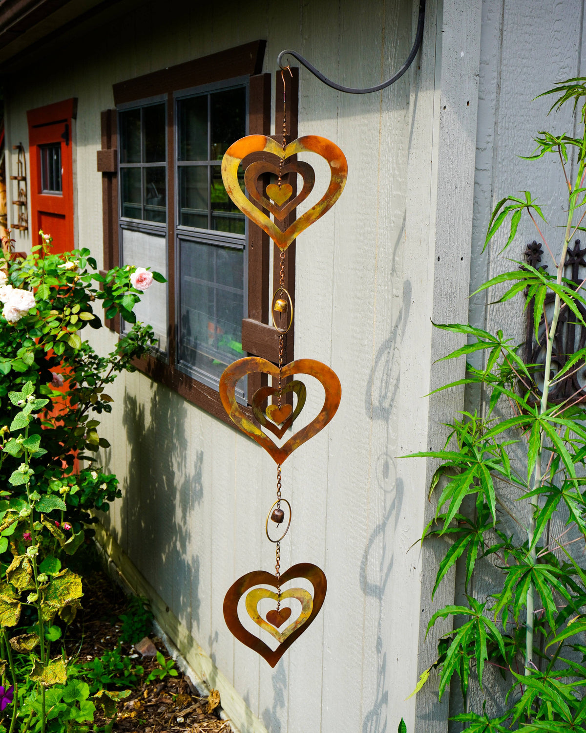 Three rustic metal heart wind chimes hang vertically on the exterior wall of a house, surrounded by green garden plants and a window.
