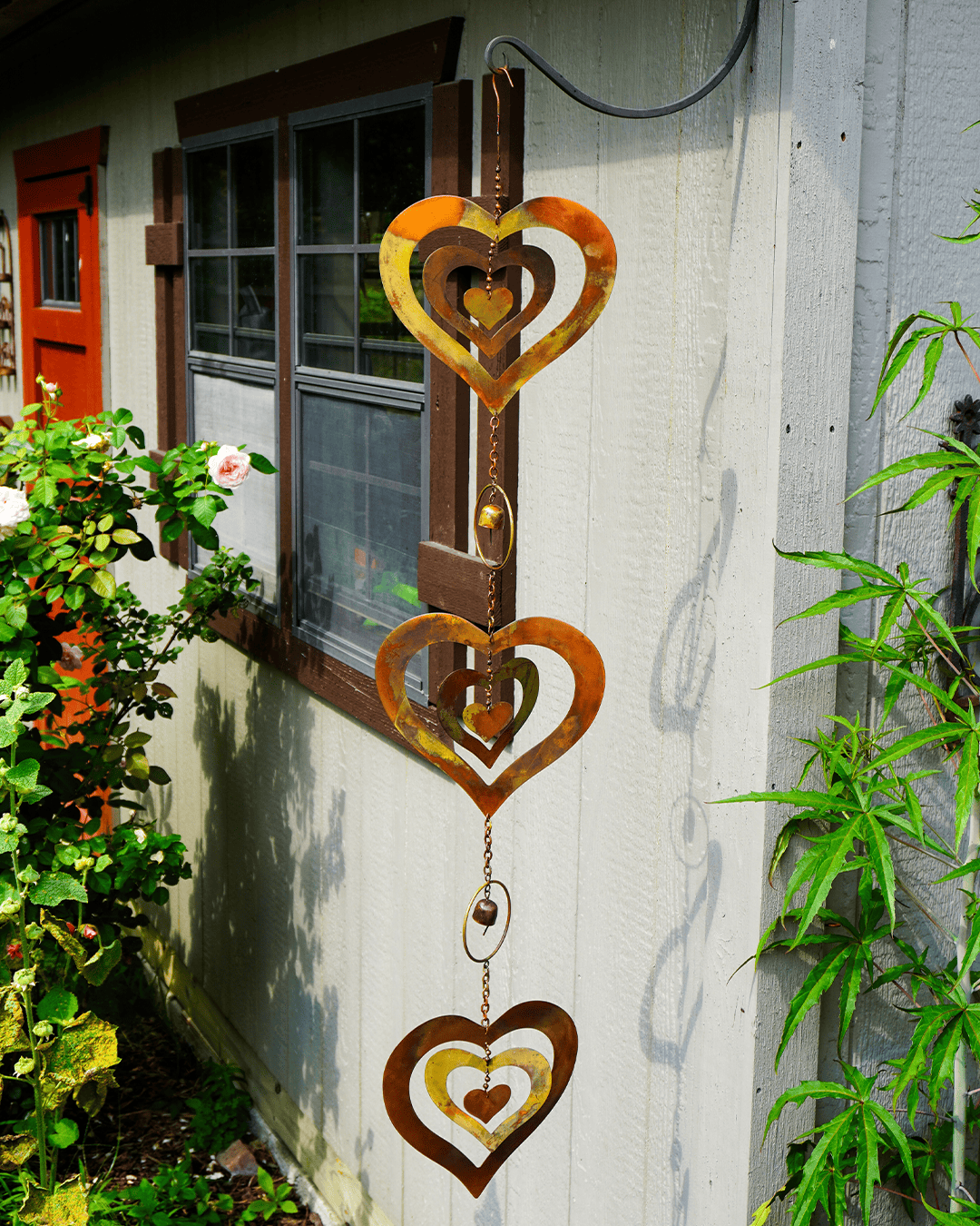 A rustic metal heart-shaped wind spinner hanging vertically on a white house exterior wall, surrounded by green plants and a window.