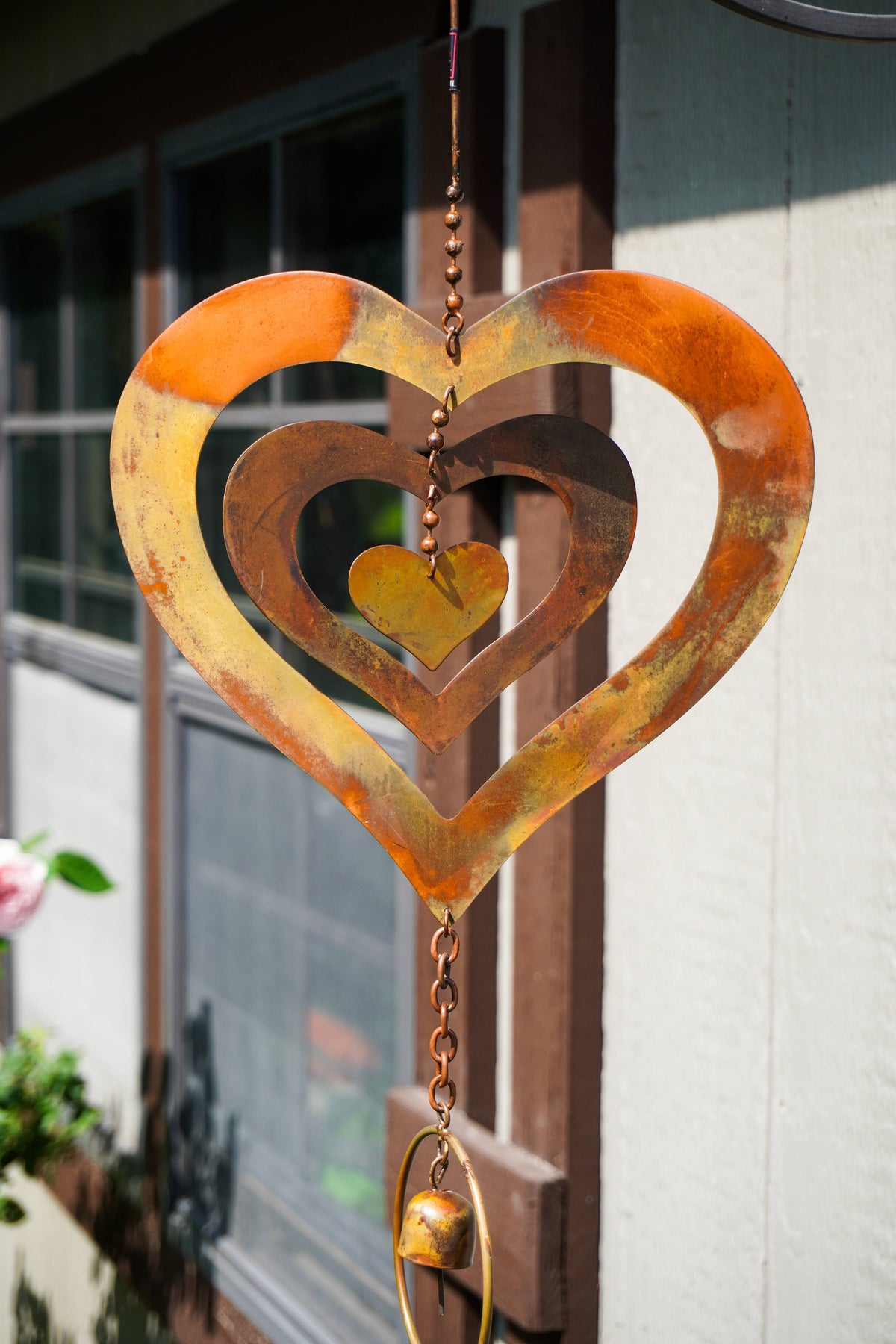 A rusted metal heart-shaped wind spinner with concentric hearts and a small hanging bell, photographed outdoors in front of a building window.
