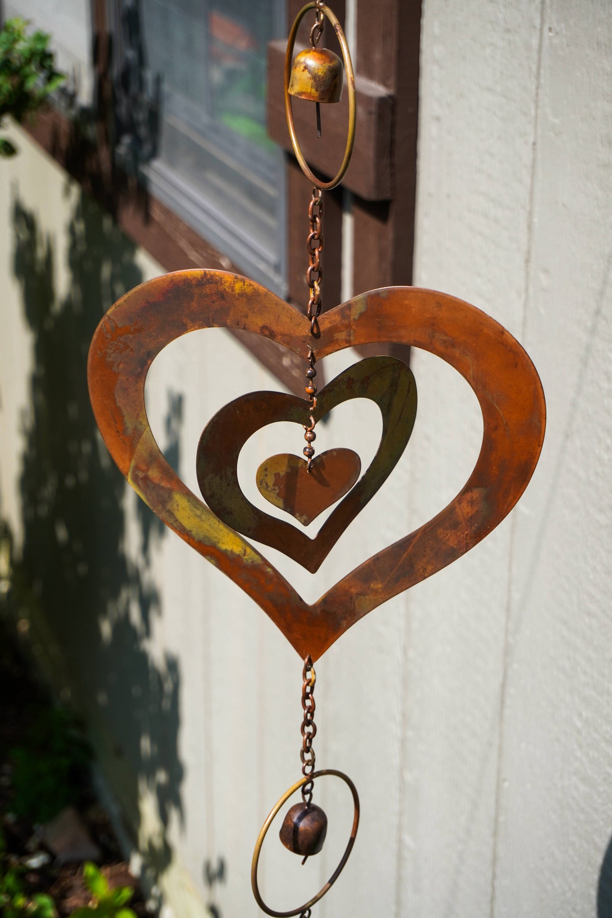 Rustic metal heart wind spinner hanging outdoors against a light-colored wall, featuring concentric heart shapes, a small hanging bell, and a dangling bead.