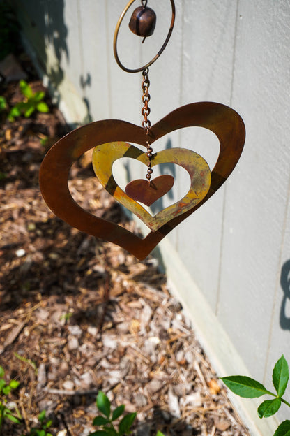 Rustic metal heart garden hanging ornament with a distressed brown finish, featuring a large outer heart, a smaller inner heart, and a central hanging chain against an outdoor backdrop.