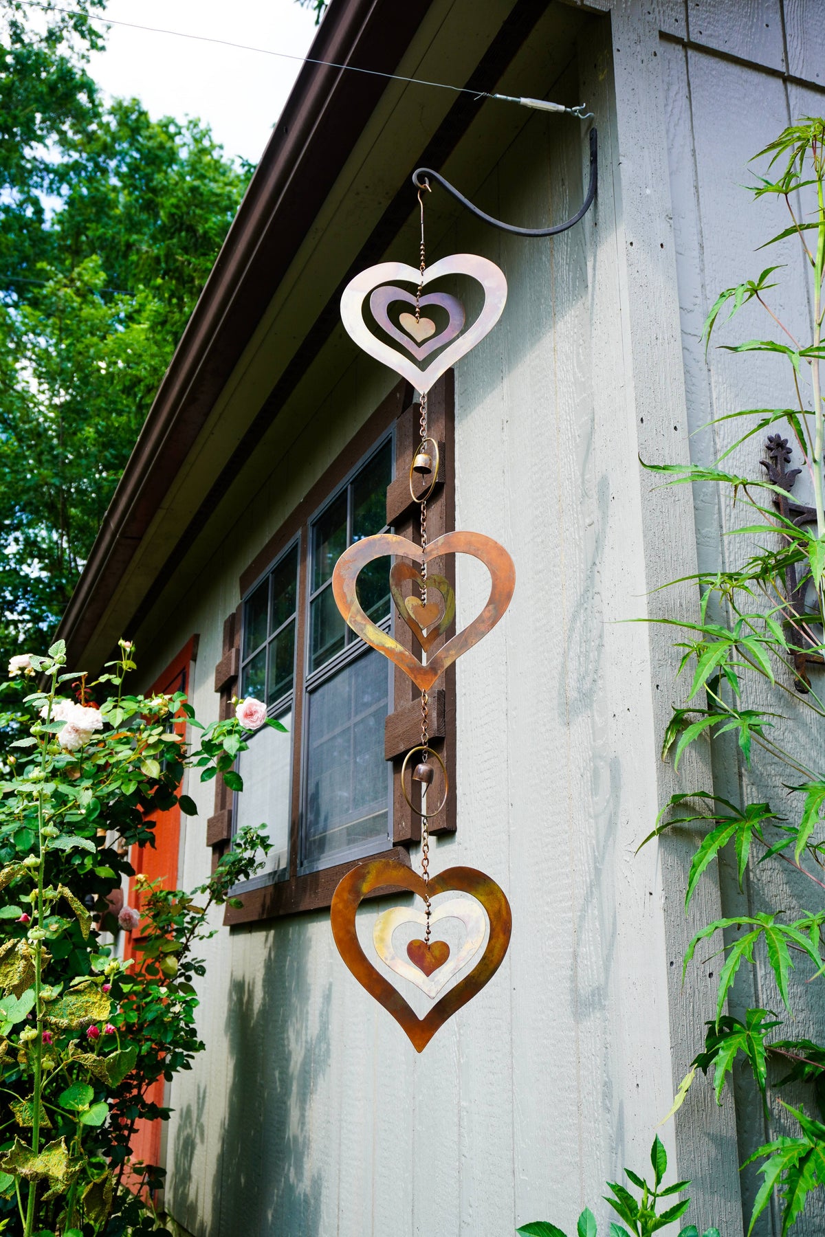 Three rustic metal heart-shaped wind spinner ornaments of varying sizes hanging vertically from a wire on the exterior wall of a house, with green garden foliage on both sides.