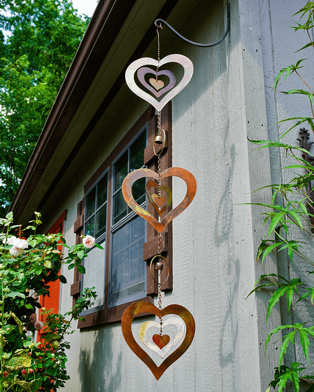 A decorative hanging wind spinner with three stacked heart shapes in white, copper, and brown, suspended outdoors against a house wall with green plants and flowers.