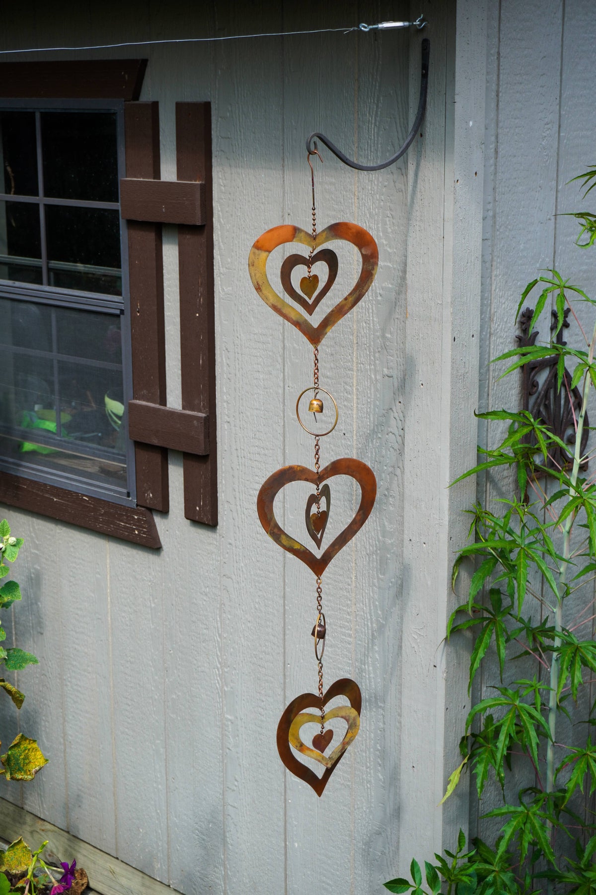 Three rusted metal heart wind chimes hang vertically on the exterior wall of a gray wooden shed, with a window on the left and green plants on the right.