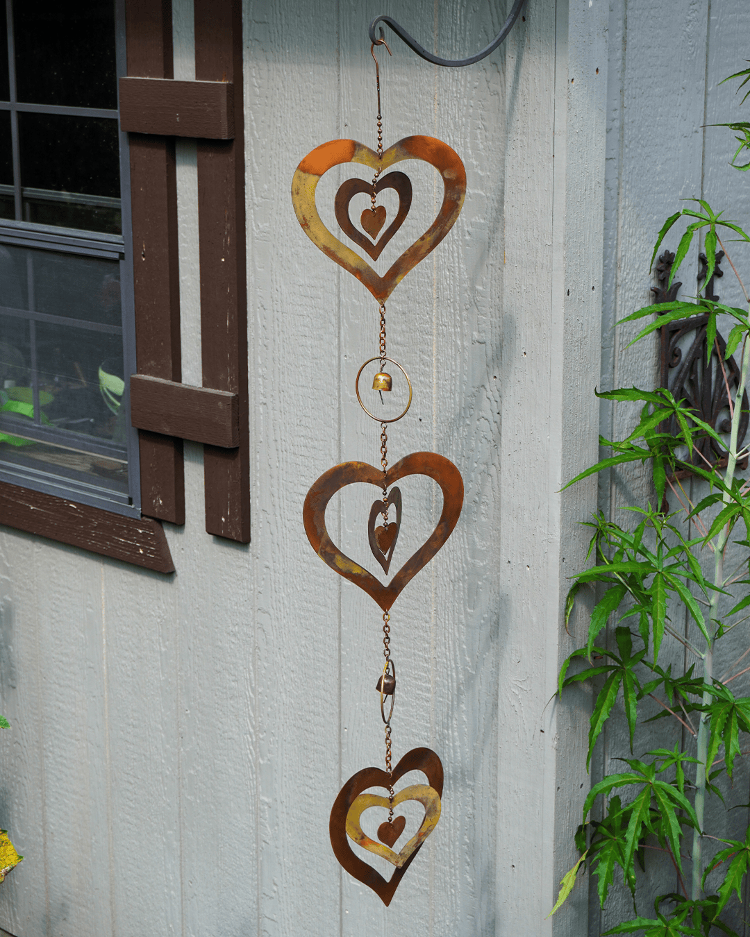 Rust-colored metal heart wind spinner hanging on a light gray exterior wall next to a window and green plants.
