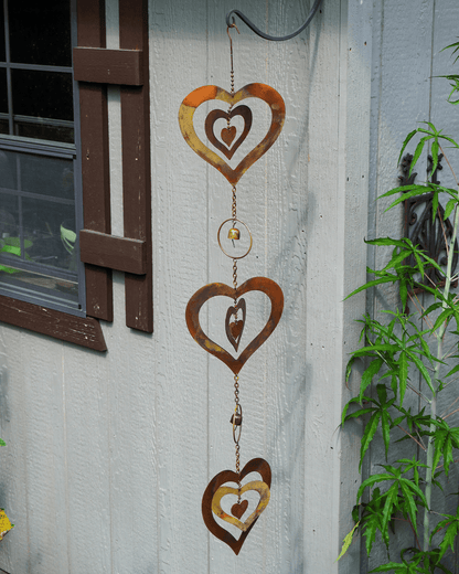 Rust-colored metal heart wind spinner hanging on a light gray exterior wall next to a window and green plants.