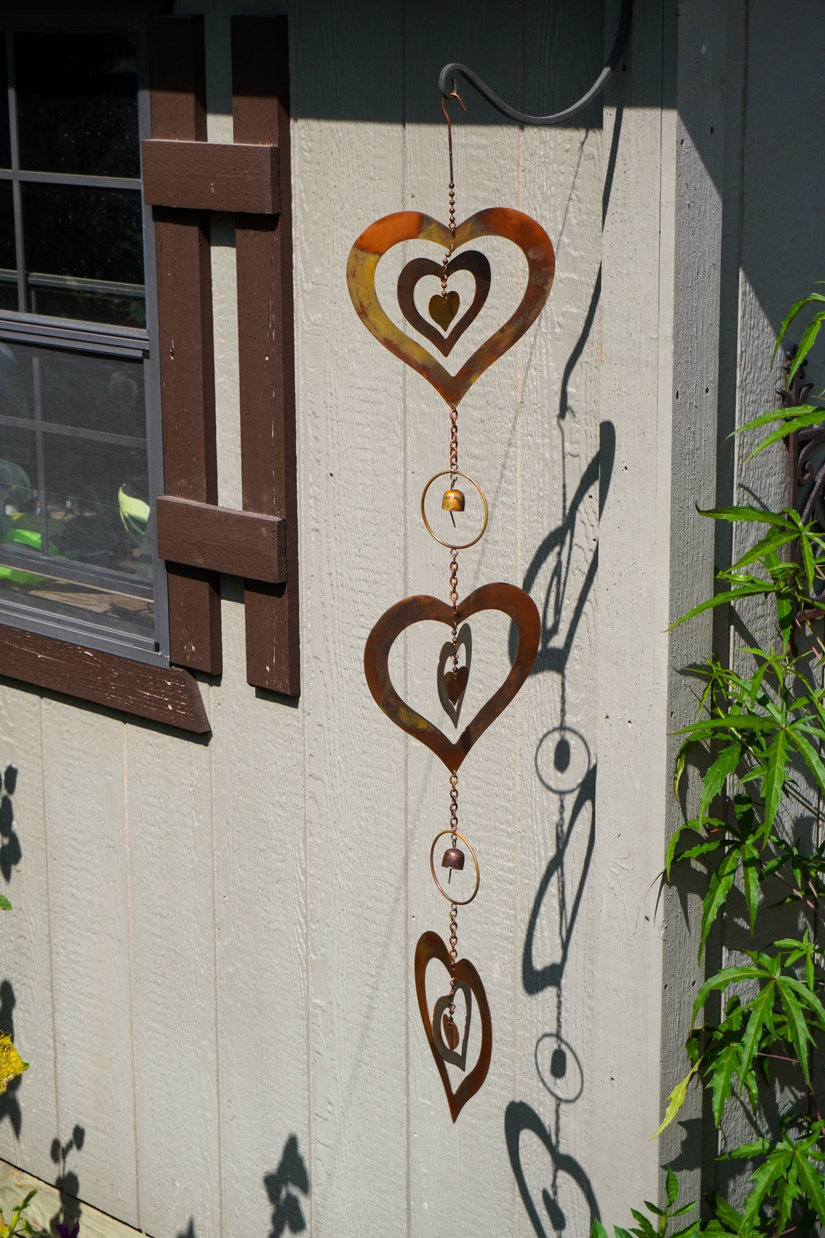 A rusted metal heart-shaped wind spinner hanging on the exterior wall of a house next to a window and green plants.