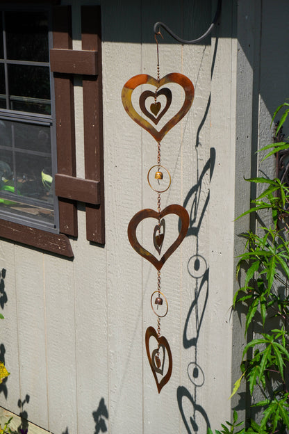 A rusted metal heart-shaped wind spinner hanging on the exterior wall of a house next to a window and green plants.