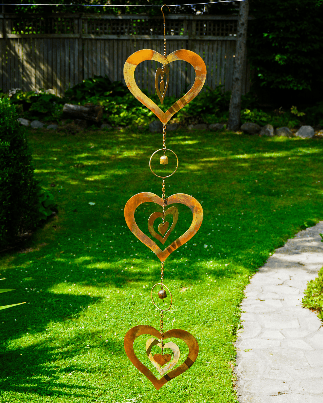 A rustic metal heart wind spinner hanging in a backyard garden. The wind spinner features three stacked heart shapes with smaller hearts inside, made of rusted orange-brown metal with gold-toned accents, suspended by chains over a green lawn.