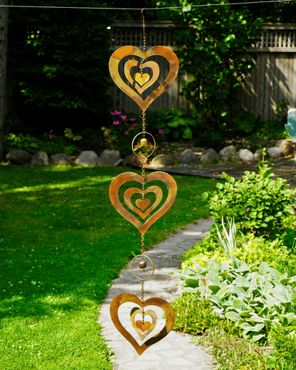 Three rusted metal heart-shaped wind chimes hang vertically on a string in a sunny backyard garden with green grass, plants, and a stone path.