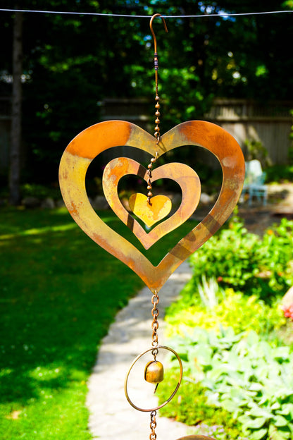 A rusted metal heart-shaped garden wind spinner hanging outdoors on a wire in a sunny garden with green grass and plants in the background.
