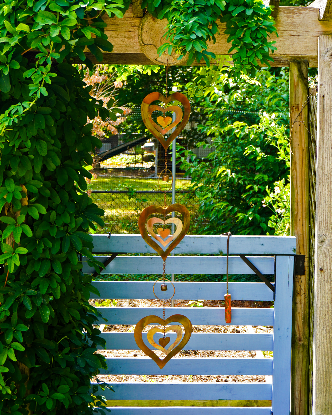 Three rustic metal heart-shaped garden wind chimes hang vertically in front of a light blue wooden gate, surrounded by lush green ivy and a garden background.