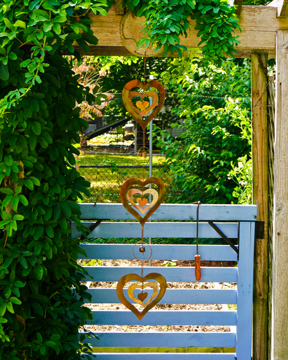 Three rustic metal heart-shaped garden wind chimes hang vertically in front of a light blue wooden gate, surrounded by lush green ivy and a garden background.
