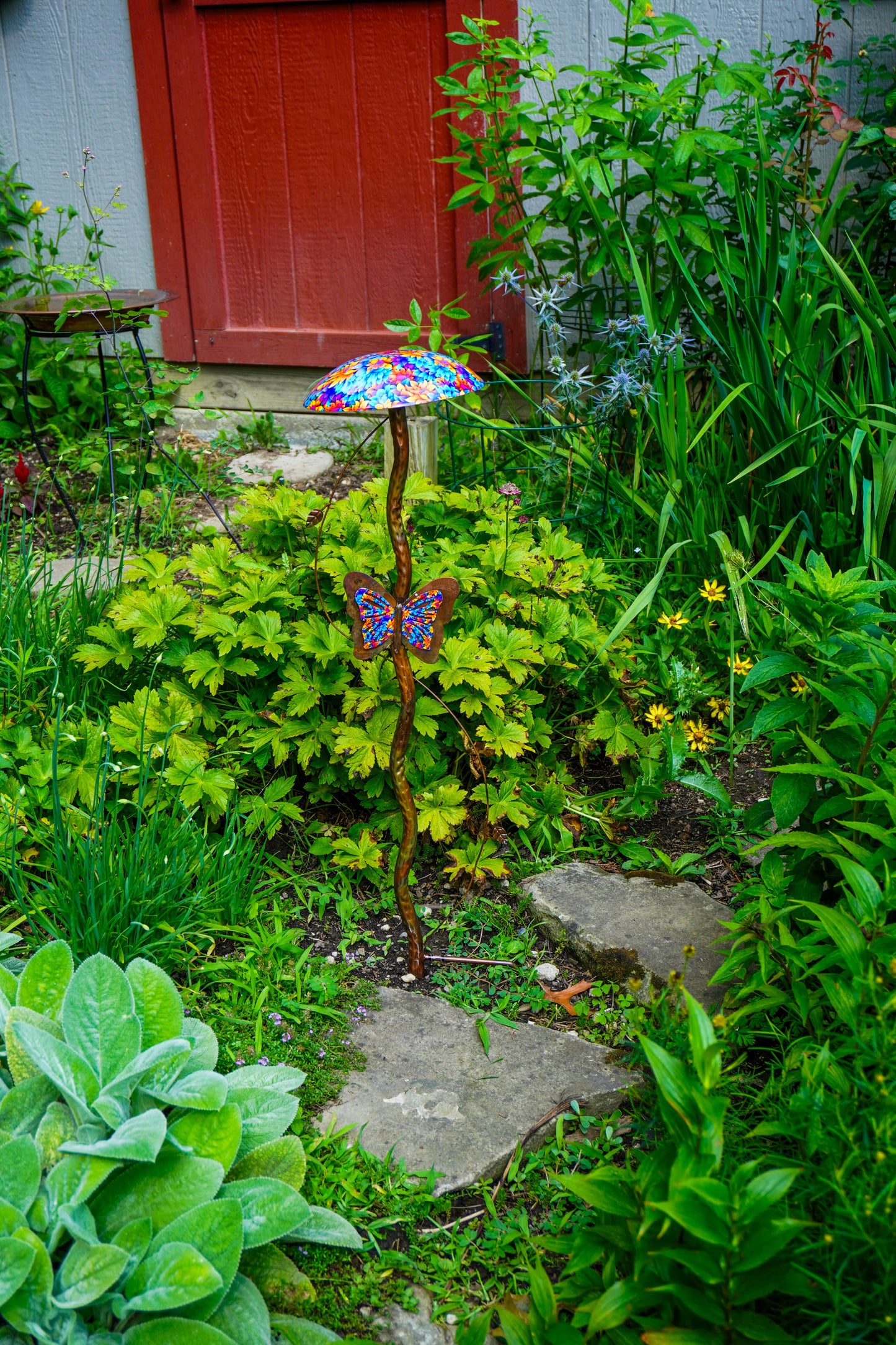 A garden stake ornament featuring a colorful stained-metal mushroom and butterfly, set in a lush green garden pathway.