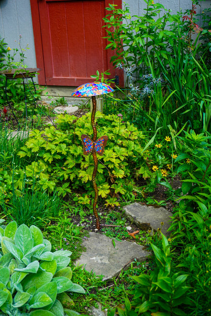 A garden stake ornament featuring a colorful stained-metal mushroom and butterfly, set in a lush green garden pathway.