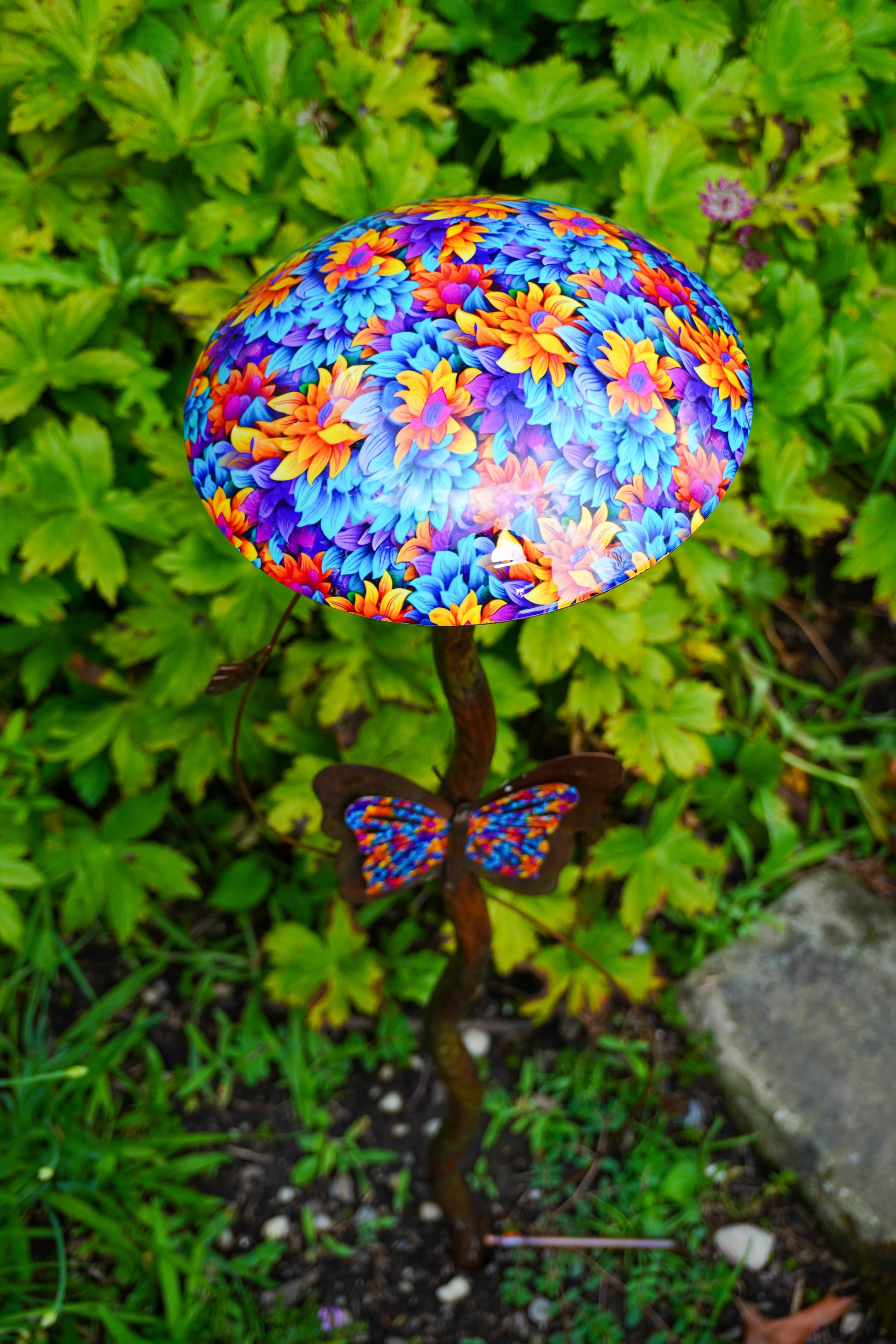 A decorative garden stake shaped like a mushroom cap with a vibrant multicolor floral pattern in blue, purple, orange, and yellow, topped with a butterfly ornament, set against a background of green garden foliage.