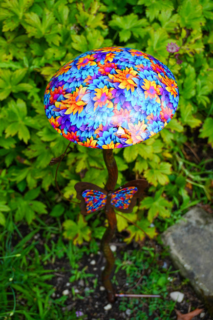 A decorative garden stake shaped like a mushroom cap with a vibrant multicolor floral pattern in blue, purple, orange, and yellow, topped with a butterfly ornament, set against a background of green garden foliage.