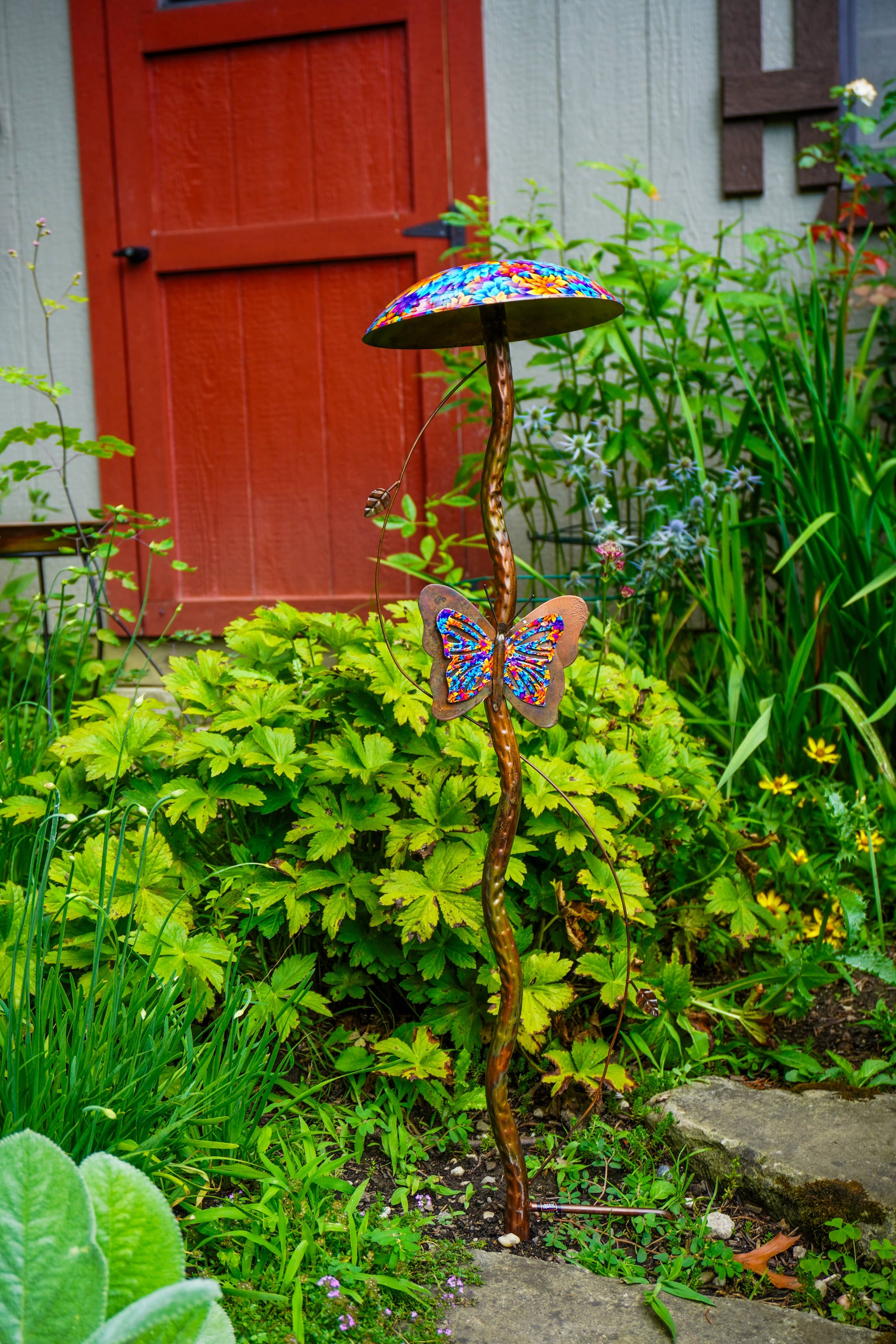 A decorative metal garden stake shaped like a mushroom cap with a colorful mosaic pattern, topped with a butterfly ornament, set in a lush garden in front of a red door.
