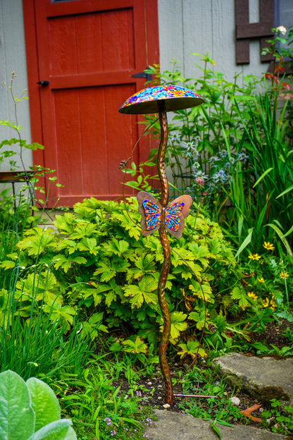 A decorative metal garden stake shaped like a mushroom cap with a colorful mosaic pattern, topped with a butterfly ornament, set in a lush garden in front of a red door.