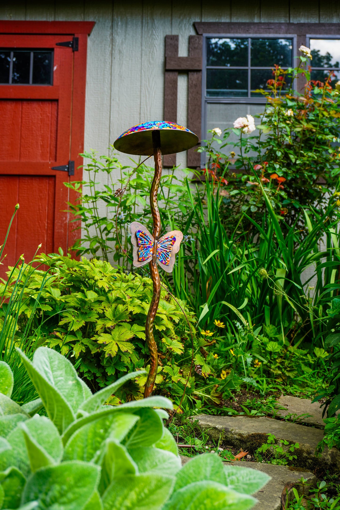 A whimsical garden stake sculpture shaped like a mushroom with a colorful patterned cap and a butterfly hanging from the stem, set in a lush green garden in front of a house with a red door.
