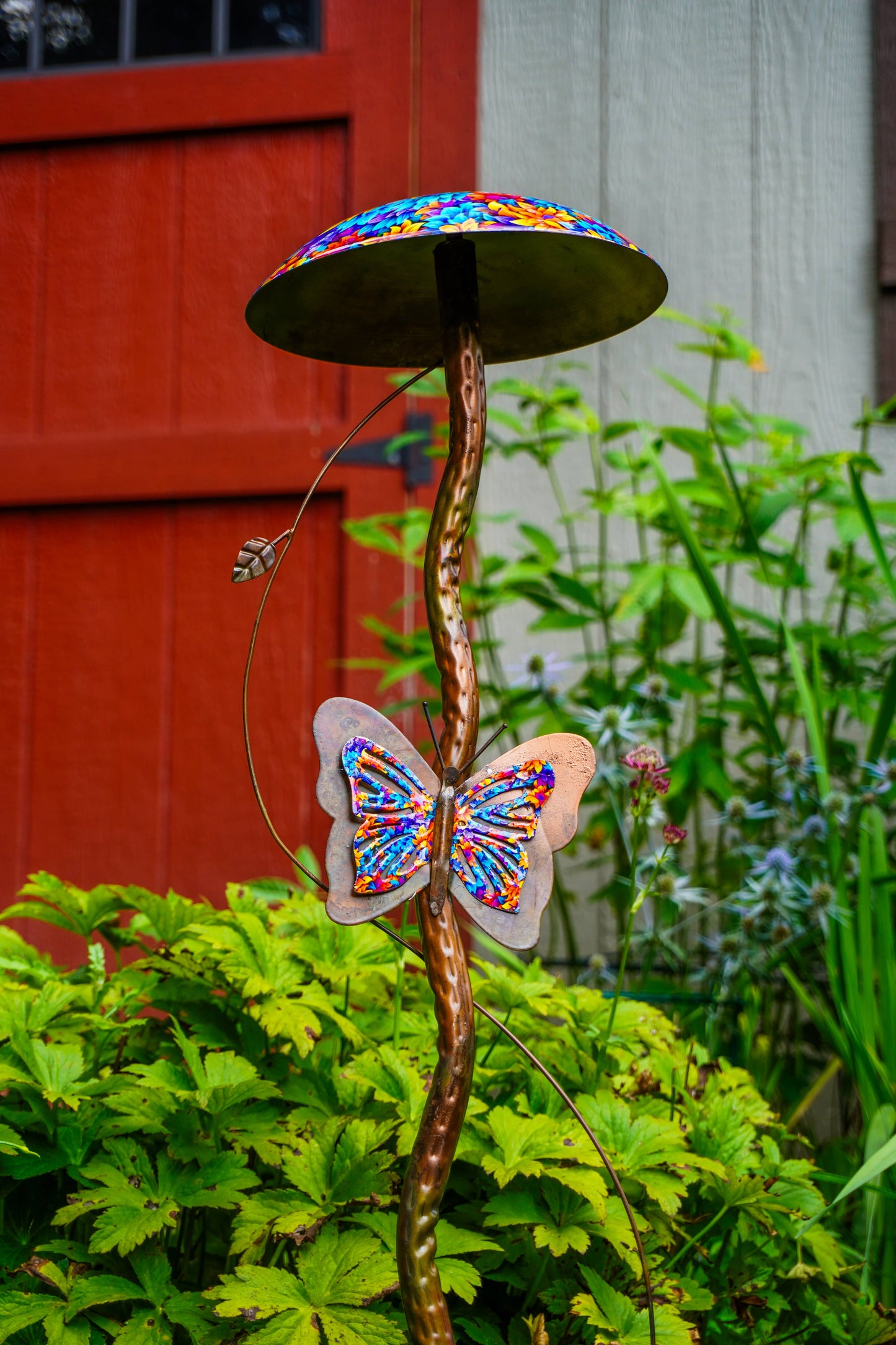 A garden sculpture shaped like a mushroom with a colorful mosaic-patterned cap and a matching butterfly attached to the stem, set in a garden with green foliage and a red wooden door in the background.