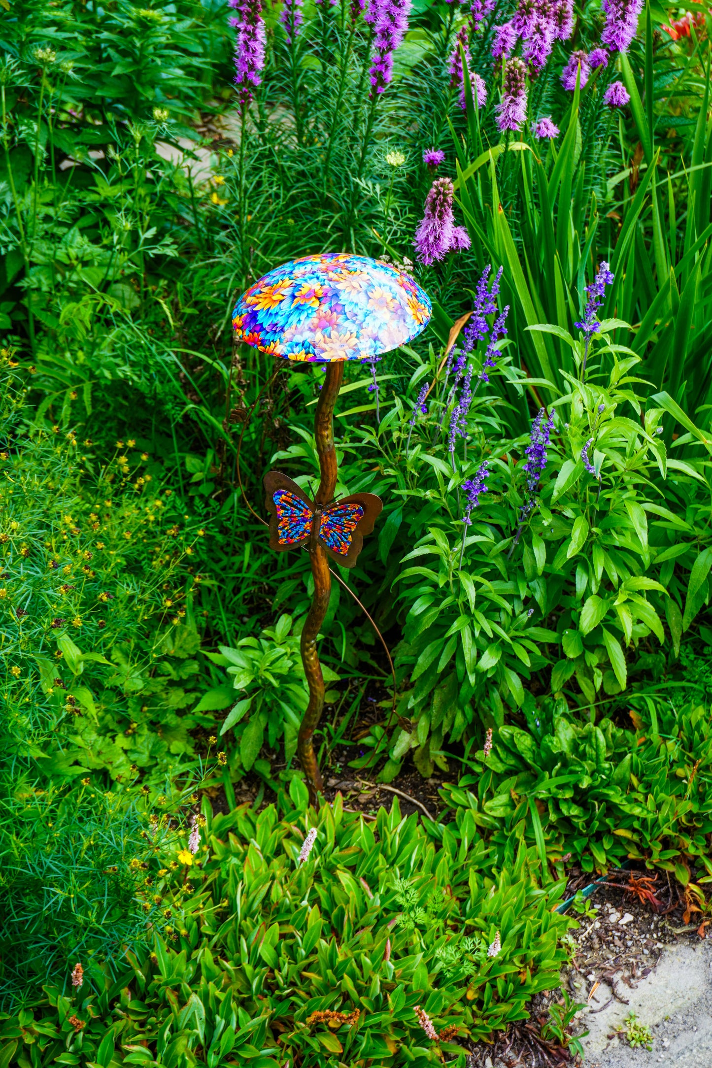 A decorative garden stake featuring a colorful, patterned mushroom cap and a butterfly-shaped metal body, surrounded by lush green foliage and purple flowers.