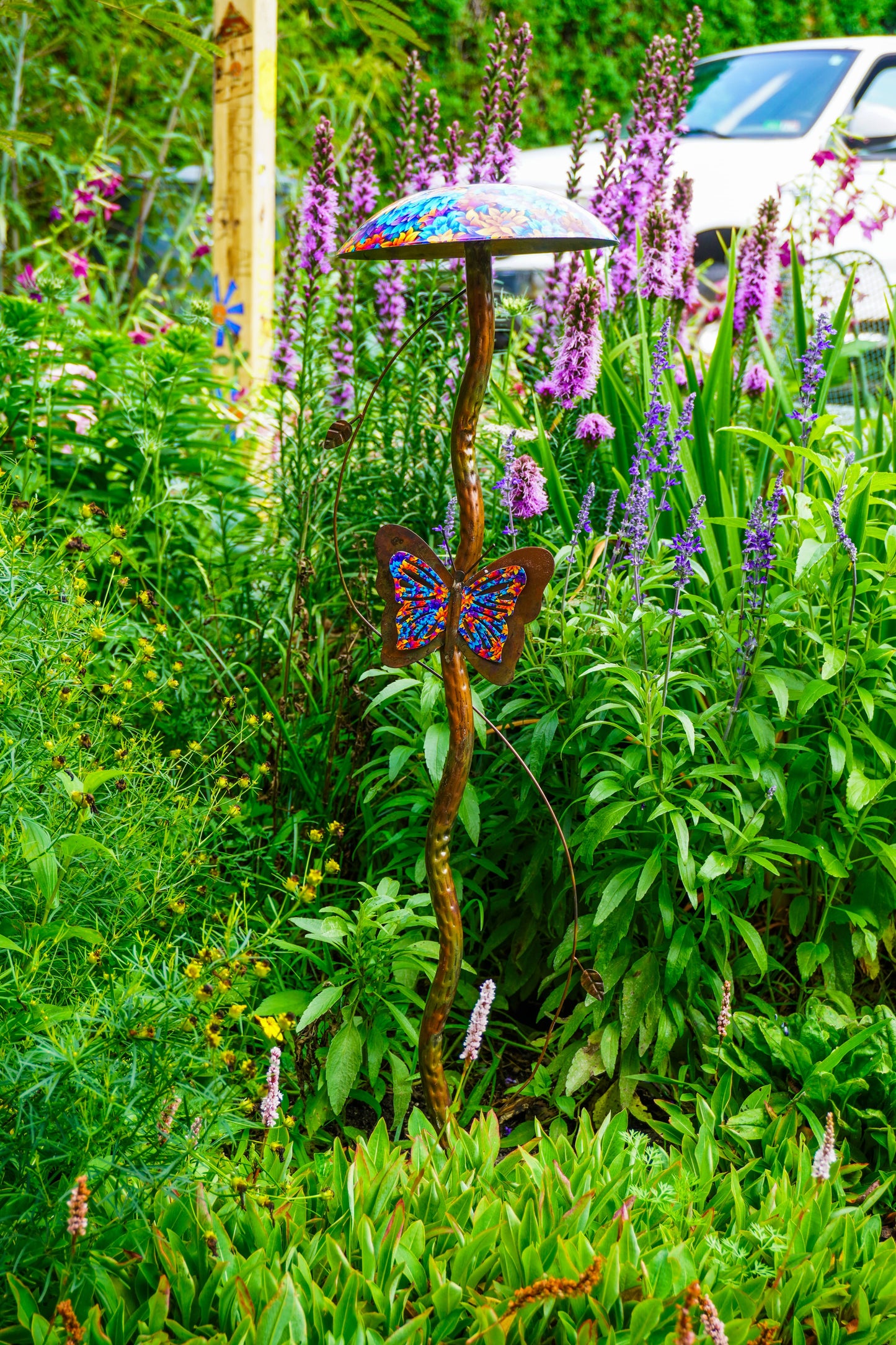 A decorative metal garden stake shaped like a mushroom with a butterfly motif, placed in a lush green garden with purple flowers. A white vehicle is partially visible in the background.