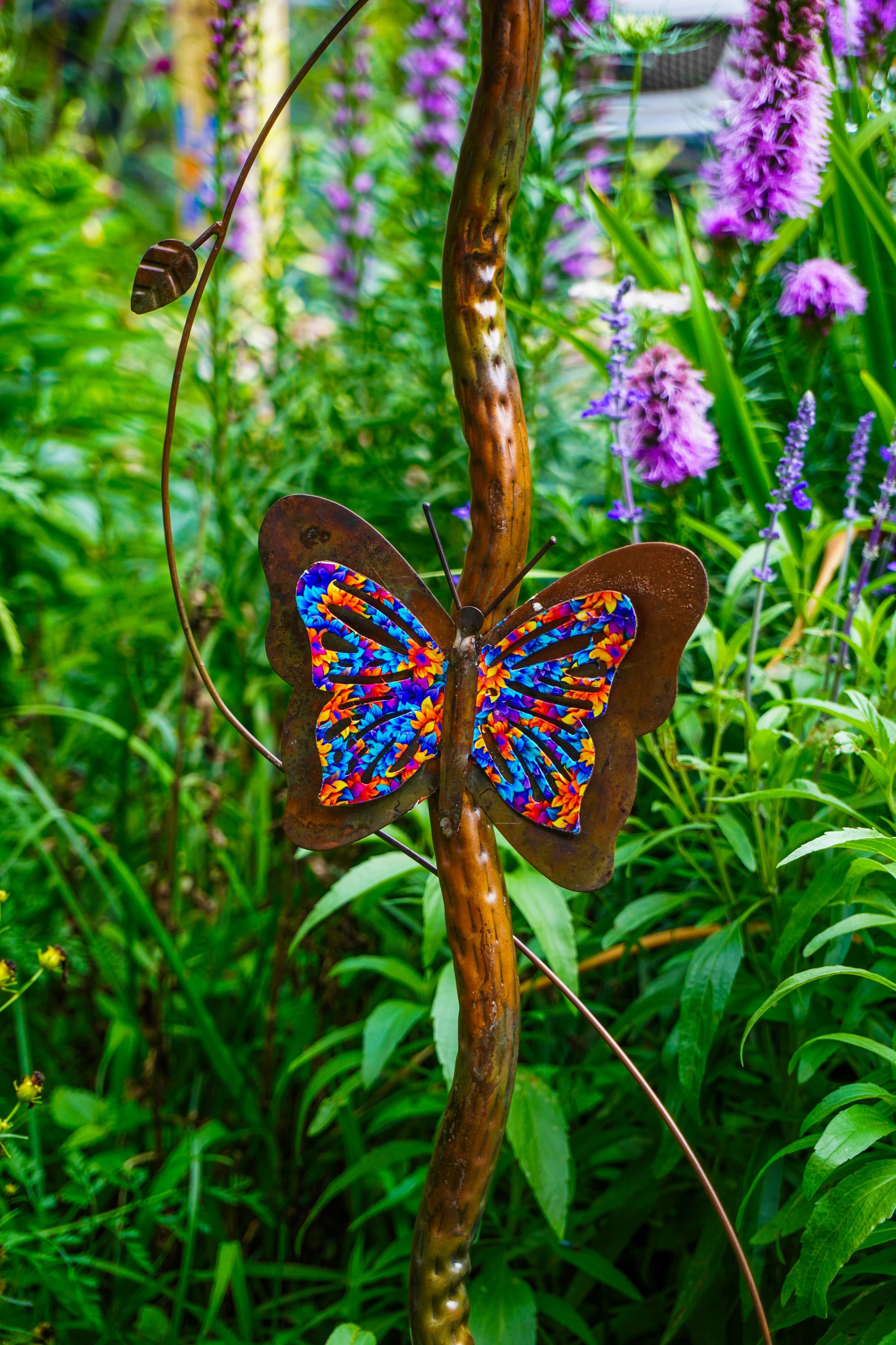 A metal garden sculpture shaped like a butterfly with rusted brown wings and a vibrant multicolor patterned center, mounted on a curved brown metal vine in a lush green garden with purple flowers.