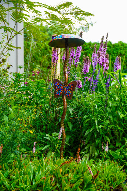 A decorative metal garden stake shaped like a mushroom cap with a butterfly mounted in the center, surrounded by lush green garden foliage and purple flowers.