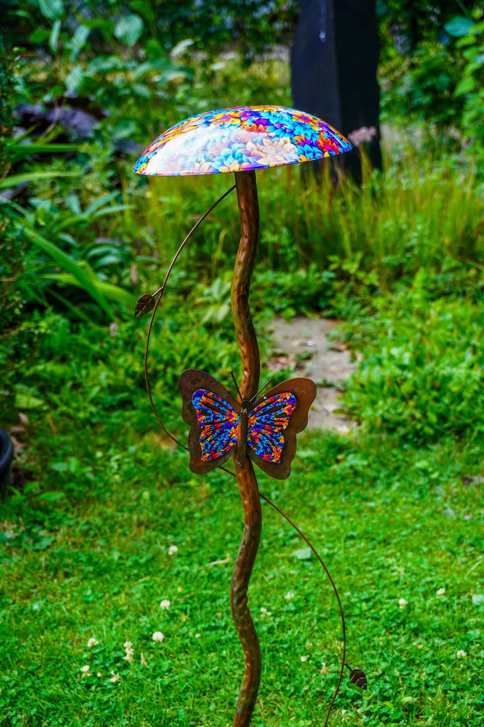 A metal garden sculpture shaped like a mushroom with a colorful stained-metal-style cap and a butterfly attached along the stem, photographed outdoors in a grassy garden.