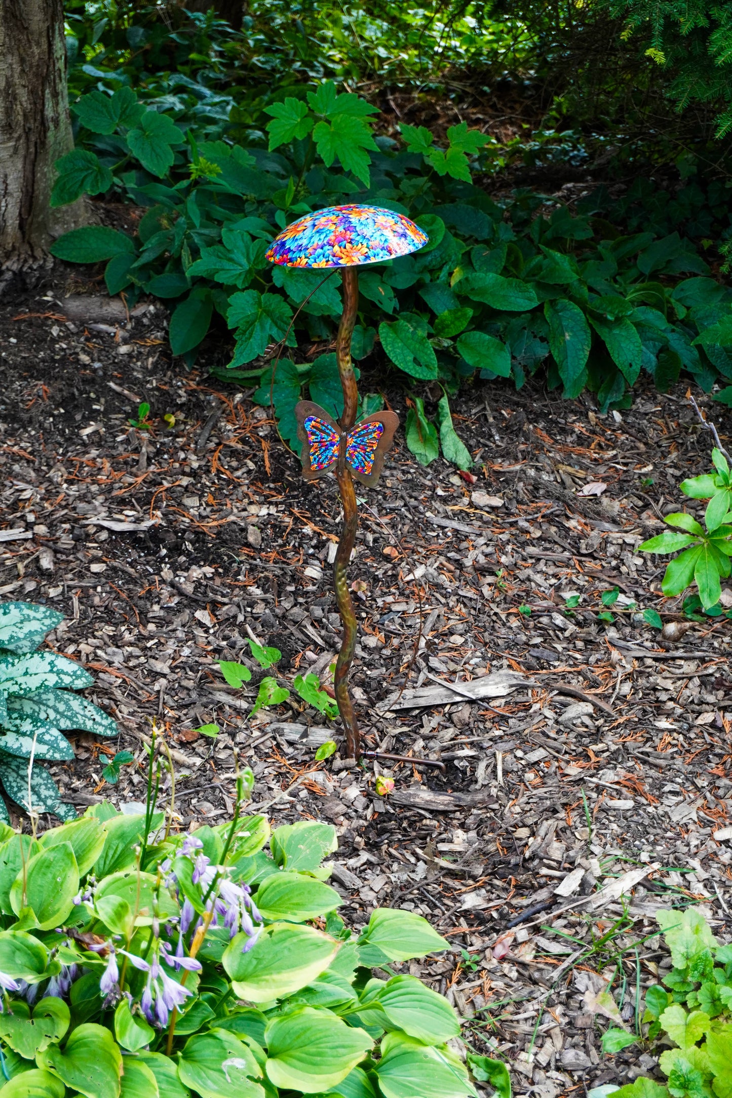 A decorative garden stake featuring a colorful mosaic mushroom cap and a butterfly-shaped ornament, set in a mulched garden bed with green plants.