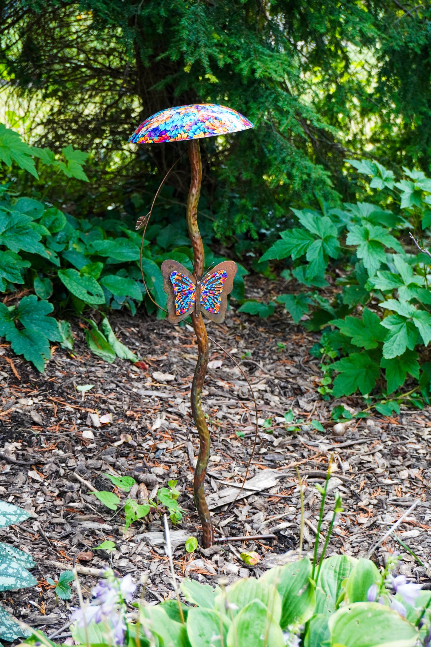 A decorative garden stake shaped like a mushroom with a colorful confetti-patterned cap and a butterfly-shaped ornament on the stem, placed in a garden bed with mulch and green plants.