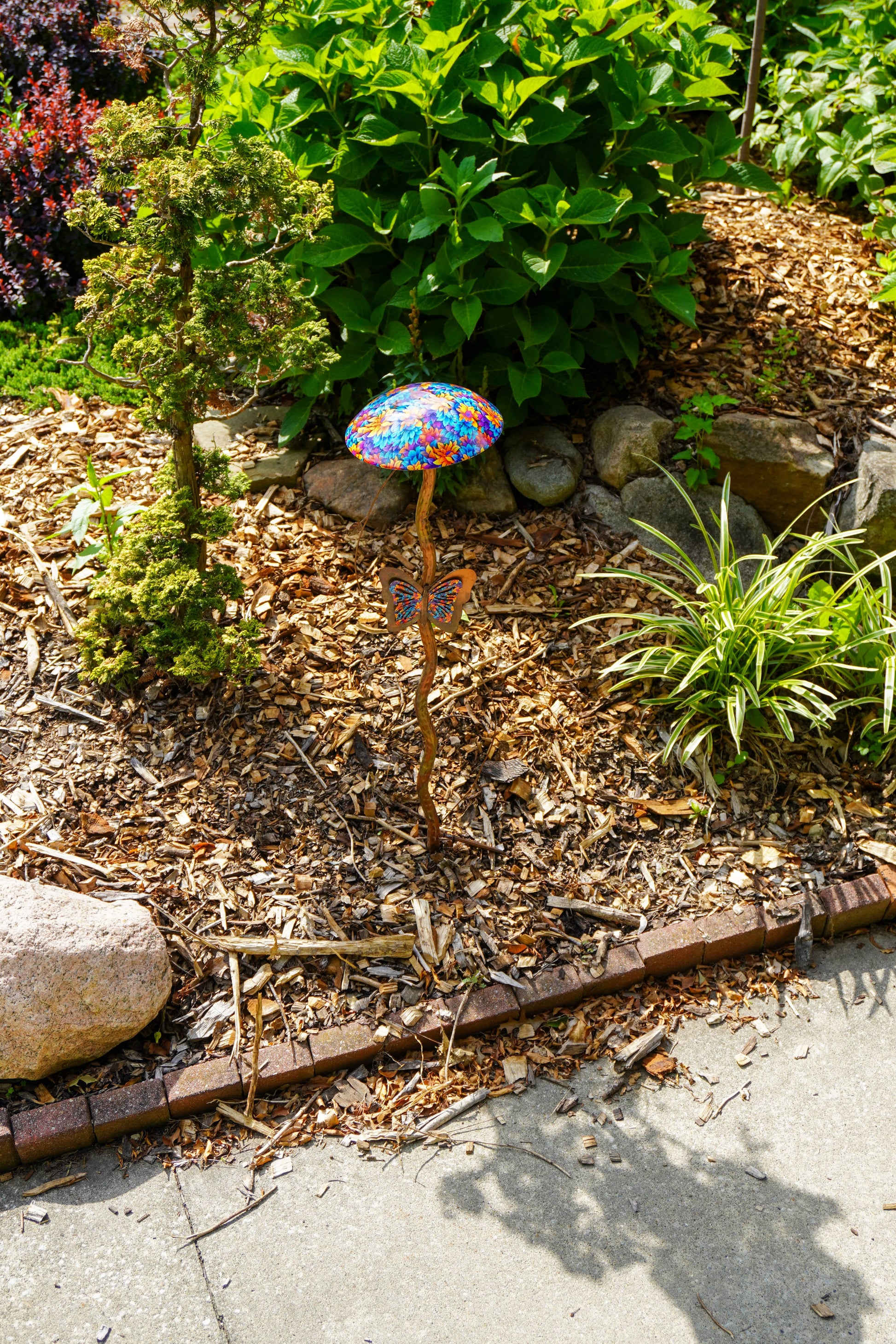 A colorful mushroom-shaped garden stake with a vibrant, multicolored patterned cap stands in a garden bed surrounded by mulch, rocks, and green plants.