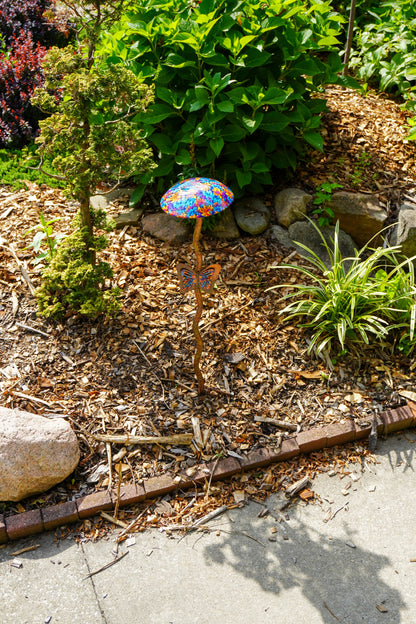 A colorful mushroom-shaped garden stake with a vibrant, multicolored patterned cap stands in a garden bed surrounded by mulch, rocks, and green plants.