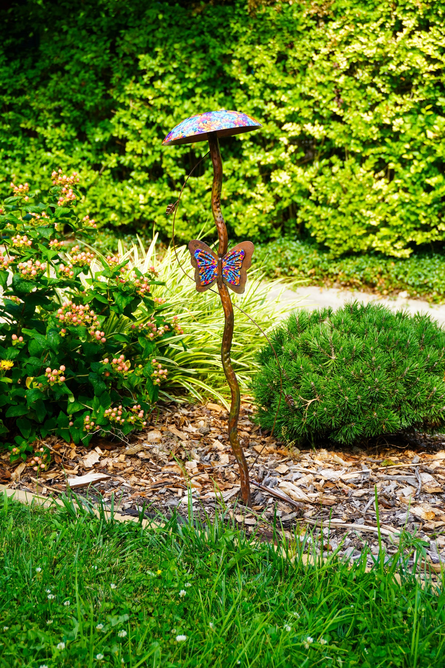 A metal garden sculpture shaped like a mushroom with a butterfly on its stem, placed in a landscaped garden bed with mulch, grass, and shrubs.