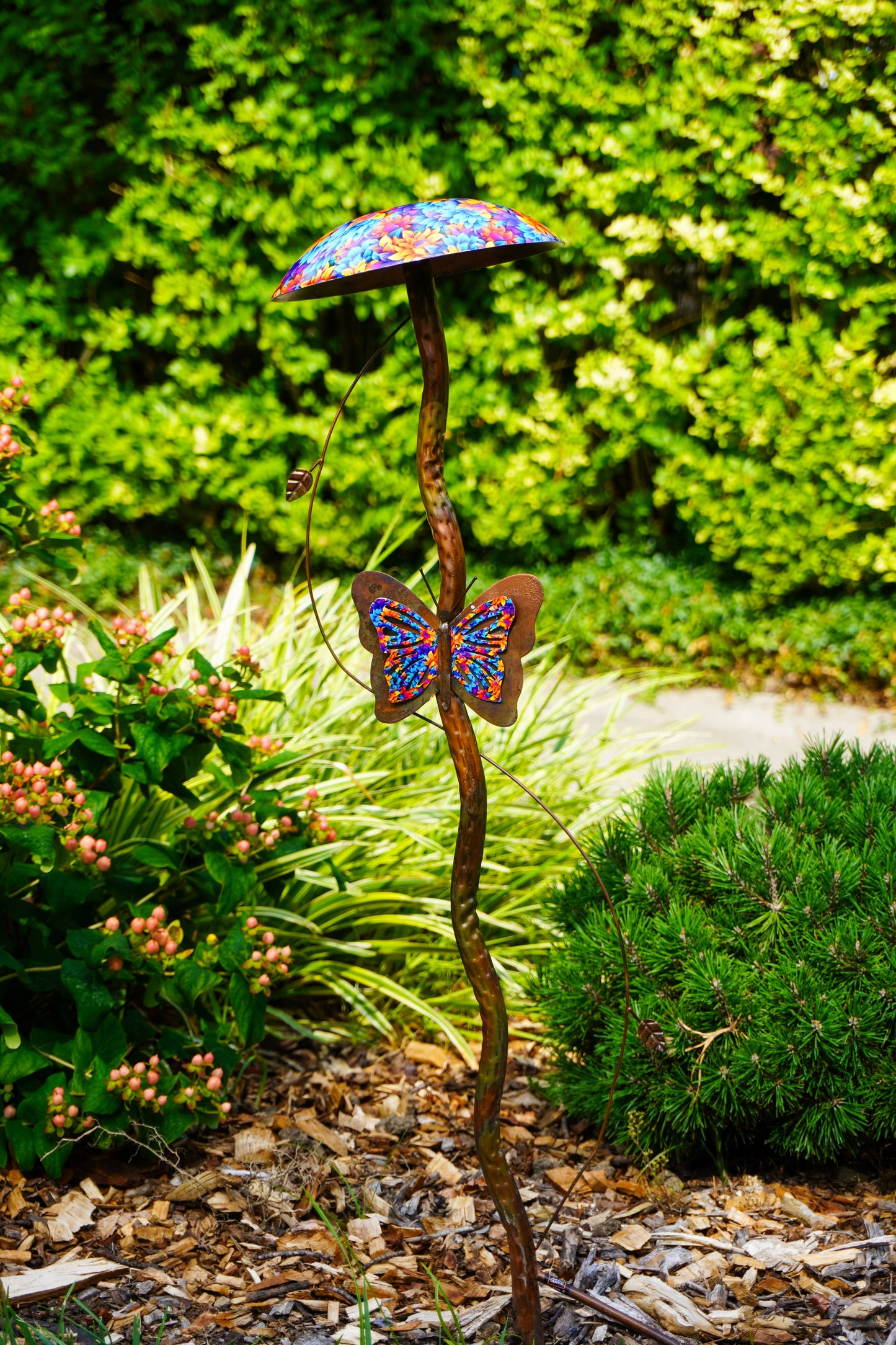 A metal garden stake sculpture shaped like a mushroom with a colorful patterned cap and a butterfly motif on the stem, set in a garden with mulch, greenery, and a pathway.