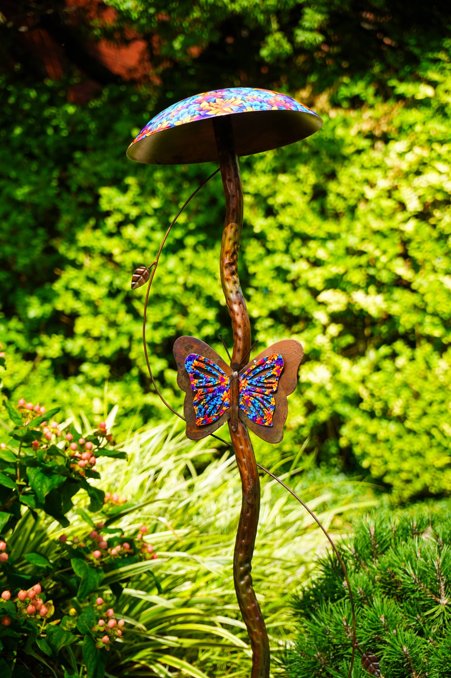 A garden metal sculpture shaped like a mushroom with a butterfly on its stem, decorated with colorful mosaic tiles, set against a lush green garden background.