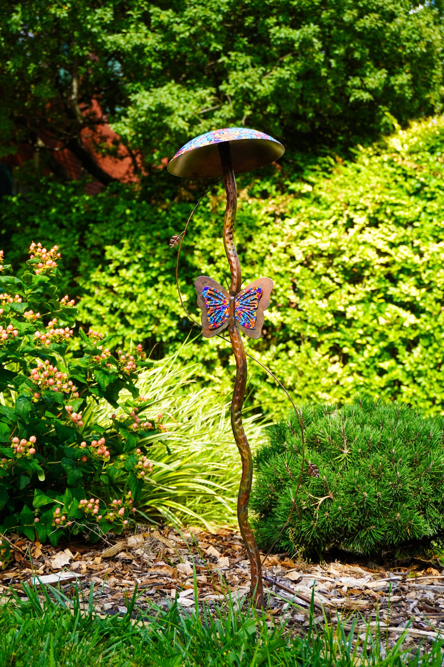 A garden metal sculpture shaped like a mushroom with a twisted brown stem and a colorful butterfly decoration on the side, set in a lush green garden.