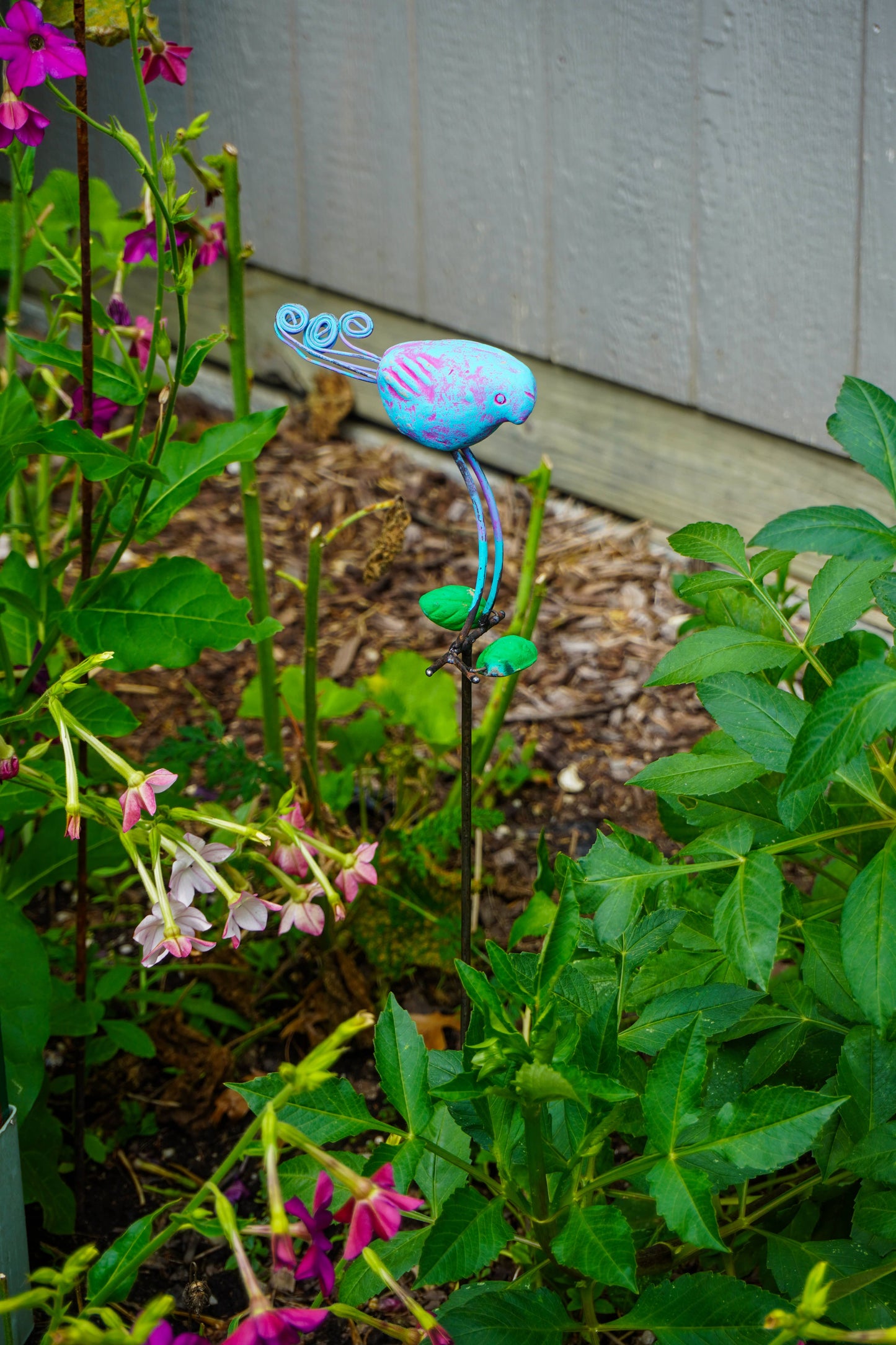 A decorative metal garden stake shaped like a small blue bird with pink accents, planted among green garden foliage and pink flowers in a garden bed.