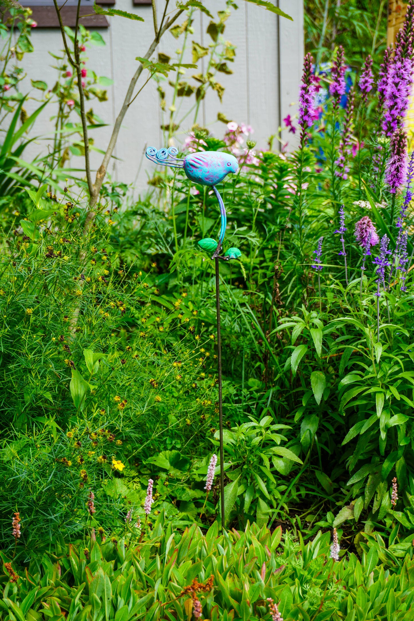 A blue metal garden stake sculpture shaped like a stylized flower or bird, placed in a lush green garden with purple flowers and foliage.