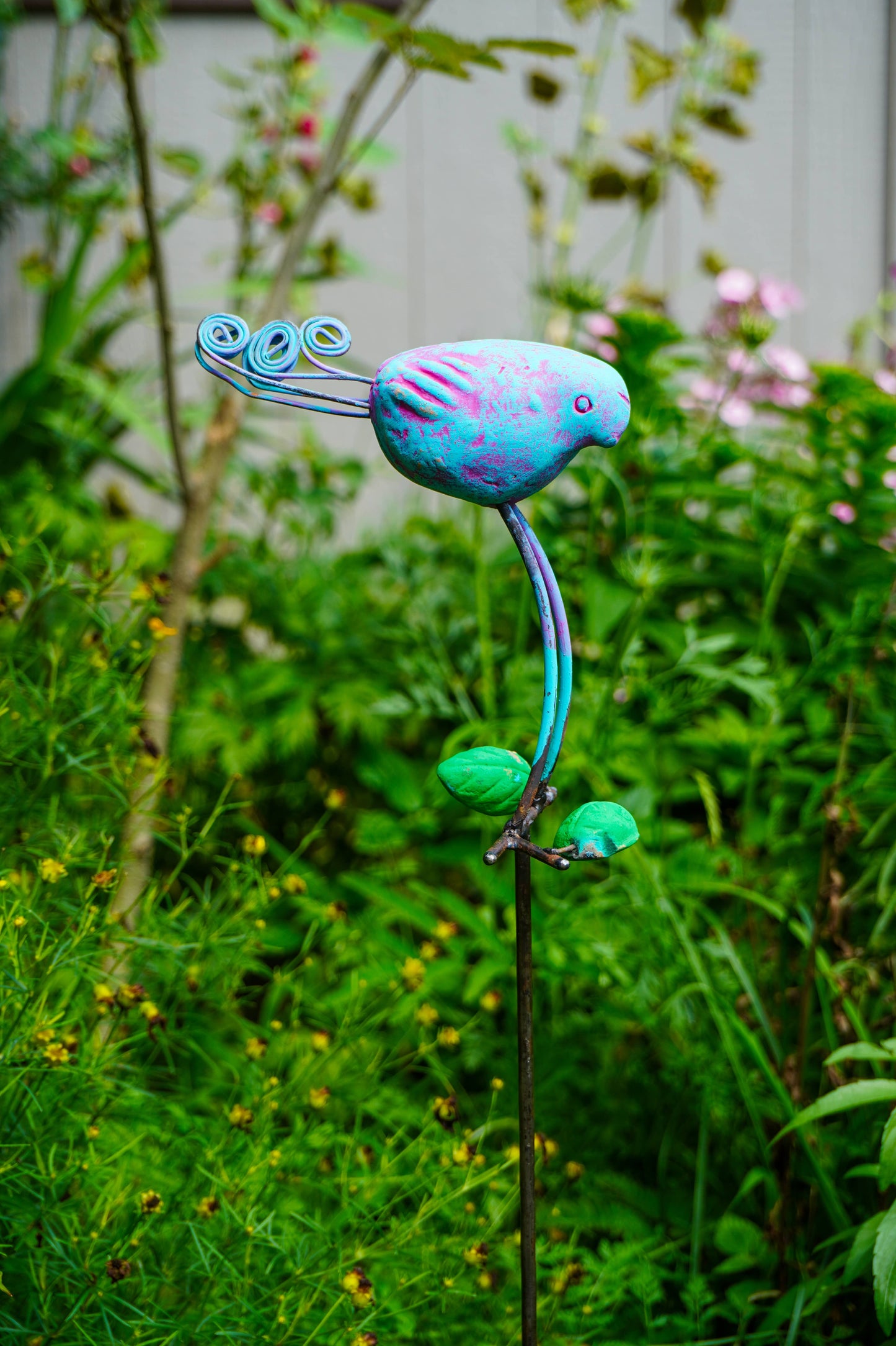 A colorful metal garden sculpture of a stylized bird perched on a stem with leaves, photographed outdoors in a garden setting with green foliage and blurred plants in the background.